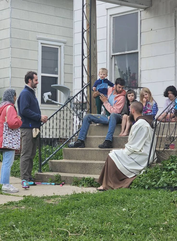 A priest sits on porch steps, talking to a family.