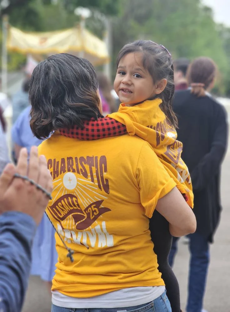 Mom with a Eucharistic shirt holds a child who is looking back at the camera