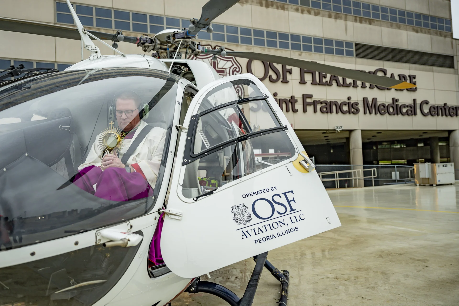 Bishop Tylka is seated inside a helicopter with the Eucharist.  The words OSF Medical Center are on the door of the helicopter.