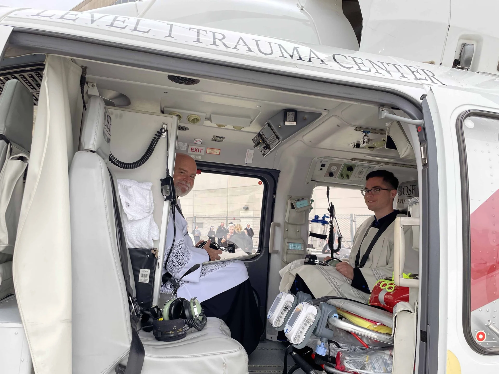 Two people in white albs sit inside a helicopter